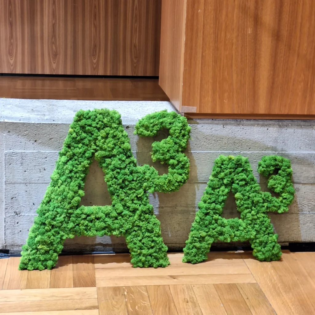 Grüne „A³“-Buchstaben aus Pflanzenmaterial stehen als Dekoration im Innenraum eines Veranstaltungsgebäudes.