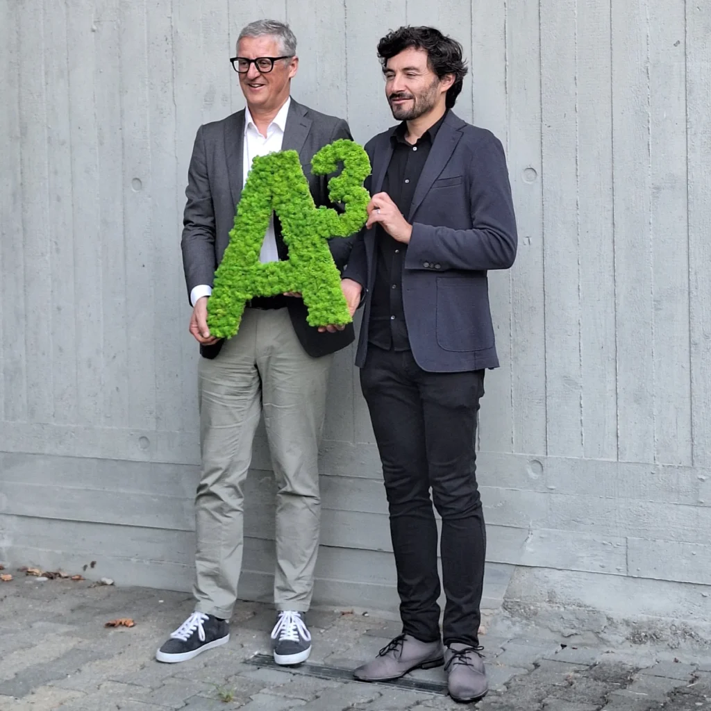 Zwei Preisträger halten ein grünes „A³“-Award-Objekt vor einer modernen Betonwand.