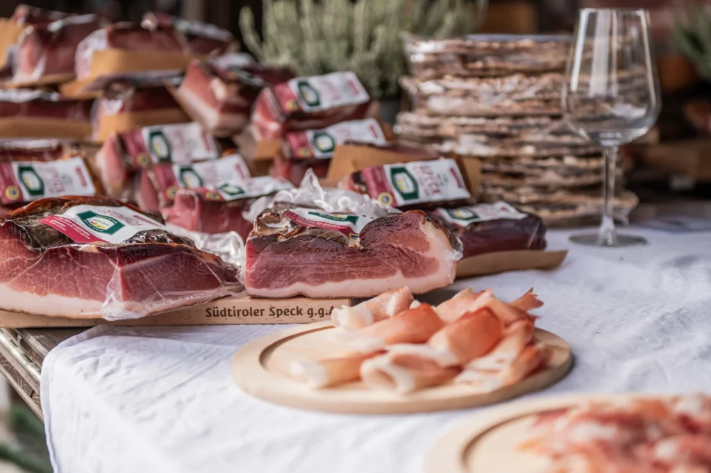 Auswahl an vakuumverpacktem Südtiroler Speck und aufgeschnittenen Speckscheiben auf einem Marktstand.