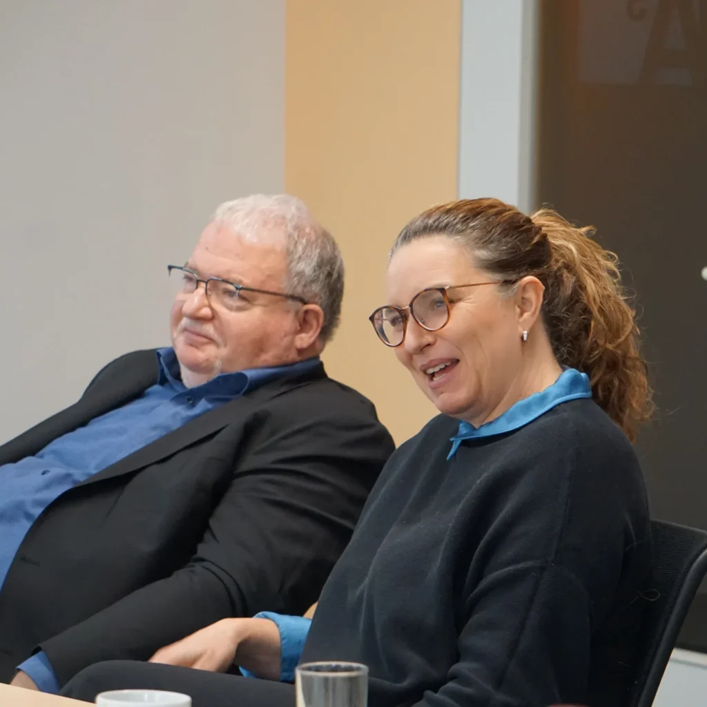 Zwei Teilnehmende sitzen an einem Tisch und hören aufmerksam einer Workshop‑Diskussion zu.